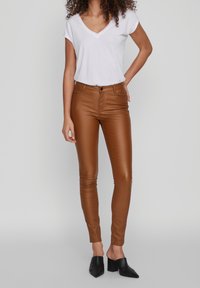 Hnědé úzké kalhoty z umělé kůže s předním zapínáním na knoflíky, doplněné bílým tričkem s výstřihem do V a černými botami na podpatku.