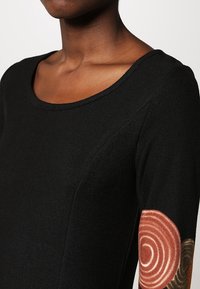 Haut noir à manches longues avec un col rond, présentant un tissu texturé et des motifs circulaires décoratifs en brun au niveau des coudes.