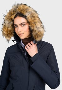 Námornícky kapucňový parka s umelým kožúškom, ktorý má zips, bočné vrecká a manžety s rebrovaním, nosený cez hnedý rolák a pruhované tričko.