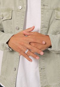Zilveren ring om vingers tegen een lichtgroene jas. Handen zijn ontspannen en tonen de natuurlijke nagelkleur. De jas heeft knoopzakken en een gestructureerde stof.