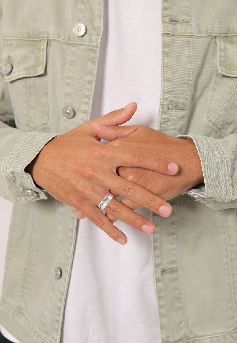 Zilveren ring om vingers tegen een lichtgroene jas. Handen zijn ontspannen en tonen de natuurlijke nagelkleur. De jas heeft knoopzakken en een gestructureerde stof.