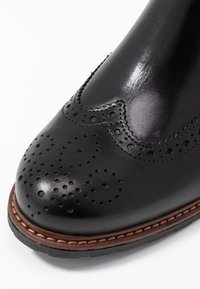 Stivale brogue in pelle nera con dettagli perforati e motivo ondulato sulla tomaia. Il bordo cucito in marrone attorno alla suola aggiunge un contrasto.