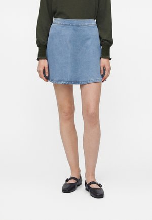 Donna che indossa una maglia verde a maniche lunghe, minigonna di jeans azzurri e ballerine nere Mary Jane, in piedi davanti a uno sfondo uniforme.