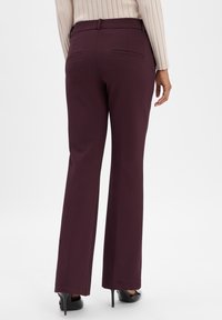 Vrouw in plomkleurige rechte broek en zwarte hoge hakken, van achteren gezien tegen een eenvoudige witte achtergrond.