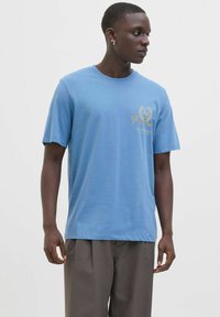 Tricou din bumbac de culoare albastru deschis, cu mâneci scurte, decolteu rotund și detaliu cu logo auriu pe piept. Asortat cu pantaloni lejeri gri.