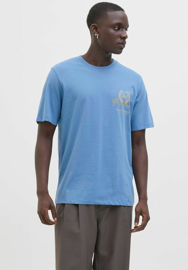 Tricou din bumbac de culoare albastru deschis, cu mâneci scurte, decolteu rotund și detaliu cu logo auriu pe piept. Asortat cu pantaloni lejeri gri.