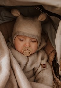 Beiger Fleece-Anzug mit Reißverschluss, einem Tierohrenhut und einem Schnuller. Eingewickelt in eine weiche Decke, die eine gemütliche Textur und neutrale Töne zeigt.