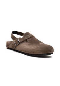 Scarpa slip-on in suede marrone con cinturino posteriore, caratterizzata da un dettaglio con fibbia argentata e una fodera in pelliccia sintetica testurizzata. Suola piatta nera.