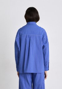 Camicia a righe blu con vestibilità rilassata, caratterizzata da un bottone nella parte posteriore del collo e maniche lunghe che arrivano fino ai polsi. Tessuto leggero.