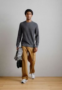 Szary sweter z długim rękawem, beżowe spodnie cargo, szaro-biała koszula w kratkę przewieszona przez ramię, białe trampki. Neutralne tło.