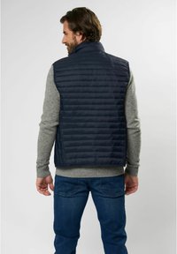 Gilet imbottito blu con motivo a righe verticali e colletto alto, indossato sopra un maglione grigio, abbinato a jeans blu. Texture morbida.