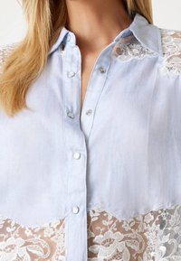 Błękitna bluzka z krótkim rękawem, z ozdobnymi srebrnymi guzikami, przezroczystymi koronkowymi detalami na ramionach oraz kwiatowymi wzorami na koronkach.