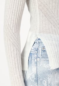 Lys grå ribbestrikket genser med en teksturert overflate, som har diagonale mønstre og en splitt på siden som avslører lys blå slangeskinn-Jeans.