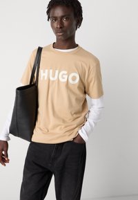Bežinis marškinėliai su ryškiu baltu "HUGO" tekstu, uždėtas ant balto ilgų rankovių marškinėlio, derinamas su juodomis džinsomis ir juoda odine rankine.