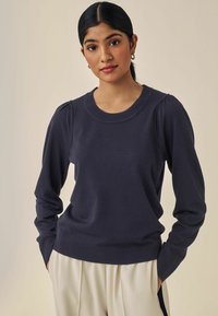 Pullover in maglina blu navy con maniche lunghe, collo e polsini a coste, arricciature sulle spalle, abbinato a pantaloni beige.