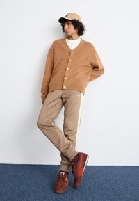 Cardigan marron tricoté avec boutons dorés, pantalon de sport beige avec une bande latérale, et baskets rouge-brun. Une casquette simple complète la tenue.