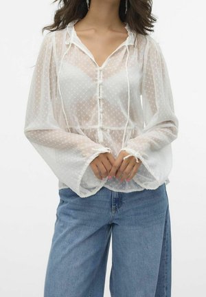 Femme portant un chemisier blanc transparent à pois avec manches longues évasées et jean bleu, les mains jointes devant.