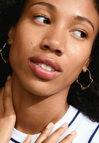 Pendientes de aro ondulados en oro, con una superficie texturizada, usados por una persona de tono de piel medio. Tiene labios brillantes y cabello natural, desordenado.