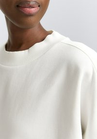 Kamera z bližnjega plaka osebe, ki nosi belo rebrasto majico s kratkimi rokavi, prikazujejoč vrat, ramo in ustnice na nevtralni ozadju.