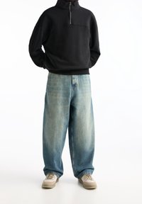 Felpa nera con zip, jeans in denim blu larghi e sneakers beige. Tessuto liscio, vestibilità comoda e texture del denim sbiadita.