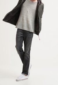 T-shirt grigio in cotone, jeans casual neri e sneakers bianche in tela. La persona indossa un giubbotto imbottito verde oliva, aperto che rivela la maglietta.
