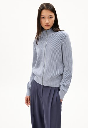 Light blue ribbed zip-up sweater with a high collar and long sleeves, paired with dark pleated trousers. Soft texture and fitted design.