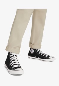 Schwarze High-Top-Canvas-Sneaker mit weißen Schnürsenkeln, Gummisohlen und einem runden Logopatch an der Seite. Getragen zu hochgekrempelten beigen Hosen.