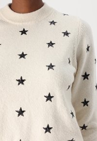 Pull en crème avec un col côtelé, ornée de motifs d'étoiles en sequins noirs répartis uniformément sur la surface du tissu. Texture douce.