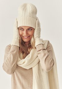 Berretto lavorato a maglia color crema con polsino a costine, guanti abbinati con motivo a trecce e una morbida sciarpa color crema drappeggiata; mostrati su uno sfondo chiaro.