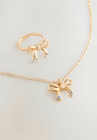 Conjunto de joyería en tono dorado que incluye un anillo en forma de lazo y un collar a juego con cadena. El collar tiene un colgante en forma de lazo con dos acentos en azul.