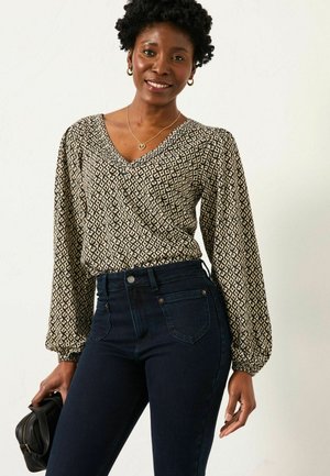 Schwarze und beige gemusterte Bluse mit V-Ausschnitt und langen Puffärmeln, kombiniert mit dunkelblauen hochtaillierten Jeans. Model hält eine schwarze Tasche.