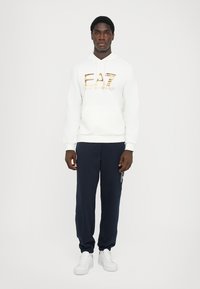 Sweatshirt branco com capuz e logo dourado EA7, emparelhado com calças de treino azul-marinho. Footwear são ténis brancos. Design minimalista, sem padrões.