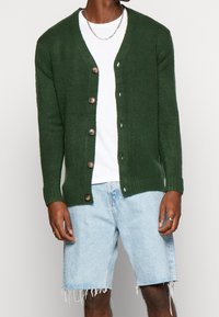 Donkergroen gebreid vest met een V-hals, knopen aan de voorkant, geribbelde manchetten, gedragen over een wit shirt en gecombineerd met lichtblauwe shorts met rafels.