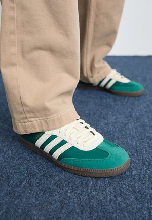 Osoba ubrana w beżowe spodnie i zielone buty Adidas Samba stoi na niebieskim dywanie.