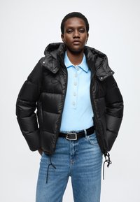 Czarna pikowana kurtka z kapturem, o błyszczącej teksturze, z regulowanym sznurkiem u dołu, noszona na jasnoniebieskiej koszulce polo. Modelka ma na sobie również dżinsy z wysokim stanem.