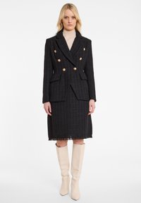 Vrouw in een zwarte dubbelbreasted blazer met gouden knopen, bijpassende knielange rok, beige coltrui en crèmekleurige kniehoge laarzen, staand op een witte achtergrond.