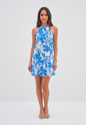 Vrouw draagt een mouwloze witte jurk met een blauw tropisch bloemenprint en naakte sandalen met hoge hakken, staand tegen een effen lichte achtergrond.