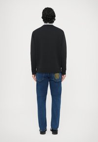 Czarna bluza z okrągłym dekoltem, zestawiona z niebieskimi dżinsami z żółtym haftem przy tylnej kieszeni. Gładkie, teksturowane tkaniny.