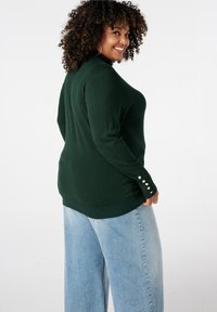 Donkergroen geribbelde trui met een hoge hals, geaccentueerd met knoopdetails op de mouwen, gecombineerd met lichtblauwe denim jeans met wijde pijpen.