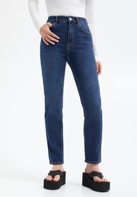 Jeans en denim bleu foncé taille haute avec une jambe droite, des coutures visibles et une fermeture par bouton à l'avant, associés à des sandales à plateforme noires épaisses.