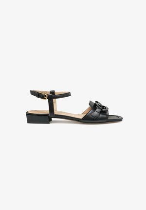 Sandalo in pelle nera con cinturino alla caviglia, fibbia dorata e dettaglio decorativo a fiocco sul davanti. Tacco basso a blocco e design a punta aperta.