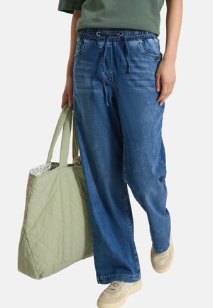 Osoba nosząca luźne niebieskie dżinsy i beżowe trampki, niosąca dużą pikowaną jasnoróżową torbę typu tote.