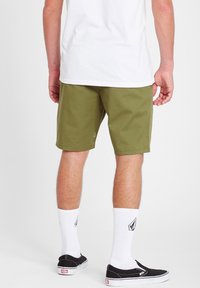 Mand iført olivengrønne shorts, hvide sokker til midt på læggen med sorte logoer, sorte slip-on sneakers og en hvid t-shirt, stående på en hvid baggrund.