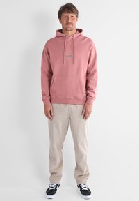 Sweat à capuche rose avec une poche kangourou, des cordons de serrage assortis et un logo bleu. Assorti à un pantalon en velours côtelé beige et des baskets noires.