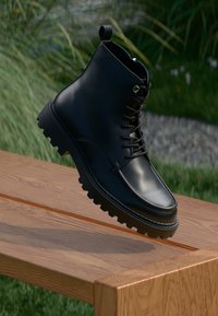 Schwarze Lederknöchelstiefel mit runder Zehenform, Schnürdesign und robuster Sohle mit tiefen Rillen, erhöht auf einer Holzoberfläche.