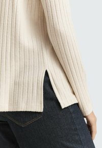 Beige geripptes Strickpullover mit seitlichen Schlitzen, kombiniert mit dunklen Jeans, die Textur und Kontrast betonen.