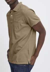 Polo shirt color canela con un bolsillo, de manga corta, con cuello acanalado, aberturas laterales y tela texturizada, combinado con jeans azul oscuro.