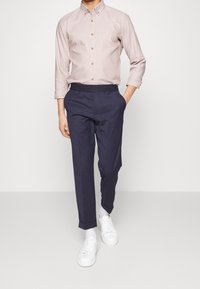 Camicia button-up rosa chiaro con maniche lunghe e bottoni marroni, abbinata a pantaloni con vita elastica blu navy e sneakers bianche.