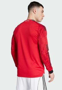 Rotes Sporttrikot mit langen Ärmeln, das strukturierte Ärmel und schwarze Streifen über den Schultern aufweist. Das Material scheint leicht und atmungsaktiv zu sein.