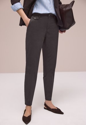 Osoba w czarnych dopasowanych spodniach, niebieskiej koszuli w paski, czarnej marynarce, trzymająca czarną skórzaną torbę i nosząca ciemne, spiczaste buty na pasku.